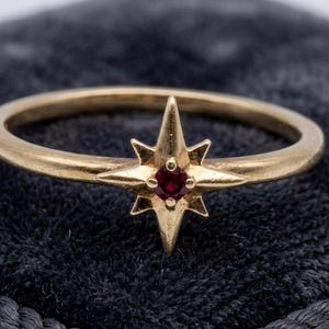 May include: A gold ring featuring a central starburst motif with a small, round, deep red gemstone set in the middle. The band is thin and smooth, and the starburst design has eight points of varying lengths, creating a celestial appearance.