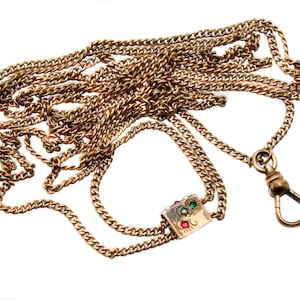 Pode incluir: Uma corrente de relógio dourada com um elemento quadrado decorativo. A corrente é composta por vários fios de pequenos segmentos ligados. O elemento quadrado é adornado com pedras preciosas vermelhas, verdes e brancas, e uma fivela é visível.