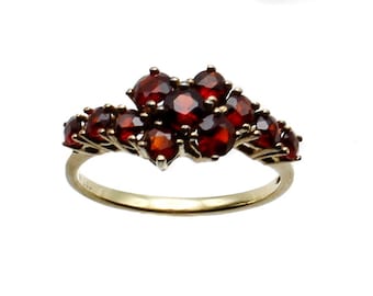 Puede incluir: Un anillo dorado con un grupo de piedras preciosas de color rojo intenso. Las piedras están dispuestas en forma de flor o de hoja, creando un diseño natural y elegante. El anillo tiene una banda pulida.