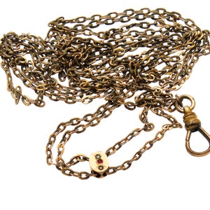 May include: Antique gold-toned watch chain with a lobster clasp and decorative oval link with red and white accents. The chain is made of small, interlocking links and is displayed against a white background.