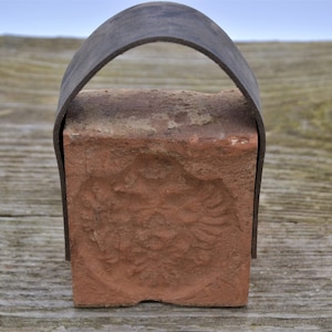 Könnte beinhalten: Ein roter Ziegel mit einem erhabenen Design auf einer Seite, gesichert mit einem schwarzen Lederriemen.