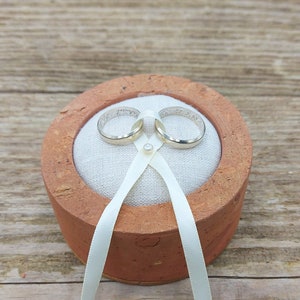 Könnte beinhalten: Zwei goldene Eheringe auf einem weißen Kissen in einer Terrakotta-Ringbox mit einem weißen Satinband.