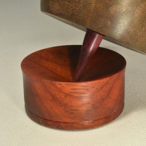 May include: A wooden spinning top with a dark brown circular body and a pointed purple tip, resting at an angle within a reddish-brown wooden base that features a concave center designed to hold the tip of the spinning top securely.