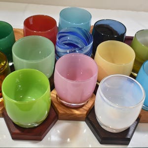 May include: A set of 12 colorful glass tumblers arranged in a hexagonal pattern on a wooden base. The tumblers are made of clear, colored, and patterned glass.