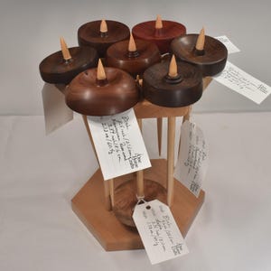 May include: A collection of seven wooden drop spindles arranged on a hexagonal wooden stand. Each spindle features a circular whorl in varying shades of brown wood with a light-colored shaft. Several spindles have white tags with handwritten text.