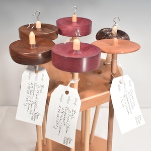 May include: A collection of six wooden drop spindles with various colored whorls, including purple, brown, and reddish-brown wood, each featuring a metal hook at the top and a white paper tag detailing specifications like 11.75 inches and 1.92 ounces.