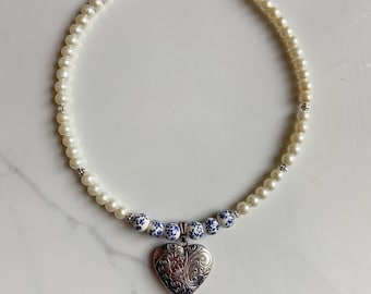 Puede incluir: Un collar de perlas blancas con un colgante de corazón de plata. El medallón tiene un diseño floral azul y blanco.