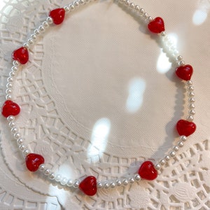 May include: A white pearl necklace with red heart-shaped beads. The necklace is on a white background.
