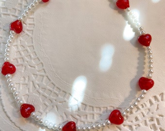 May include: A white pearl necklace with red heart-shaped beads. The necklace is on a white background.