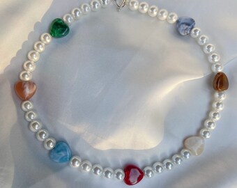 Puede incluir: Un collar de perlas blancas con un cierre plateado y cuentas de colores en forma de corazón. Las cuentas son en tonos de verde, azul, rojo, marrón y blanco.