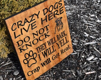 May include: A wooden sign with black lettering that reads "Crazy Dogs Live Here Do Not Knock They Will Bark I Will Yell Crap Will Get Real."
