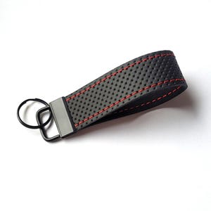 Könnte beinhalten: Schwarzer Leder-Schlüsselanhänger mit roten Nähten und einem schwarzen Metallschlüsselring. Das Leder hat ein strukturiertes, perforiertes Design.
