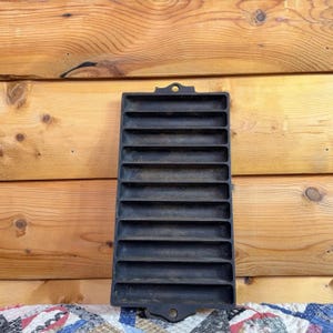 May include: A black cast iron corn stick pan featuring eleven long, narrow, horizontal indentations for baking. The rectangular metal mold has a small circular hole at the top and bottom center for hanging, with a textured, dark, matte finish.