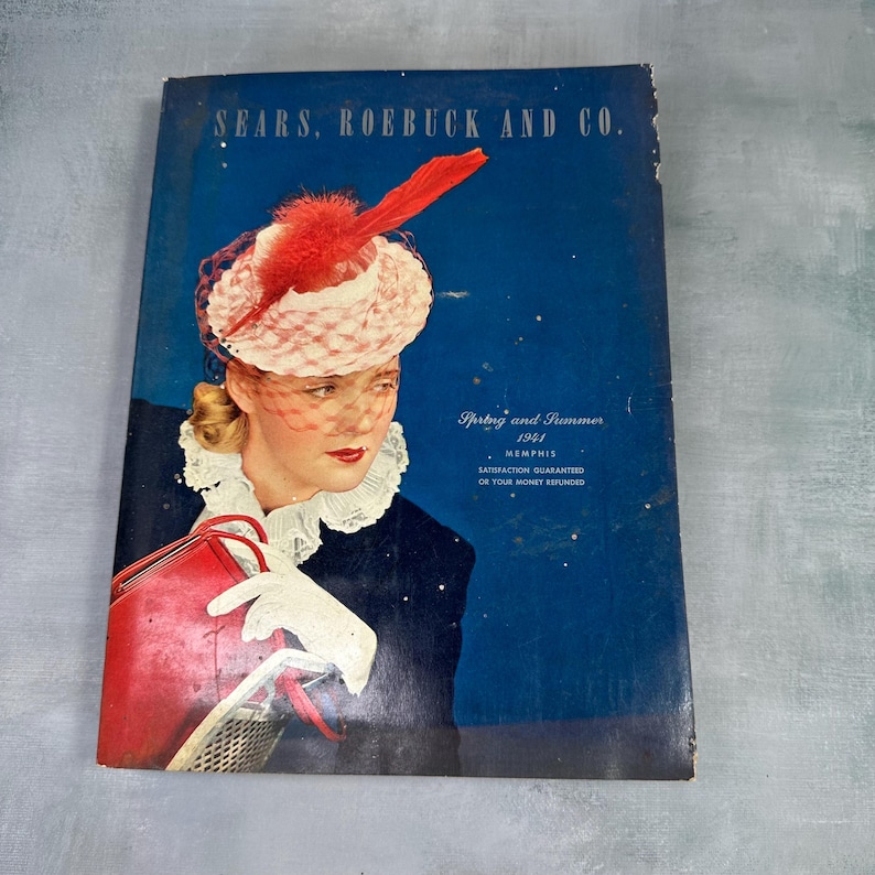 May include: A vintage Sears, Roebuck and Co. catalog cover from 1941. The cover features a woman wearing a white and red hat with a red feather, a white lace collar, and white gloves. She is holding a red purse. The background is a dark blue.