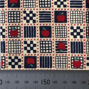 May include: A beige fabric with a repeating pattern of red apples, blue squares, and white squares. The fabric has a red and white checkered border.
