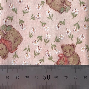 May include: Pink fabric with a repeating pattern of small white flowers and brown teddy bears. The fabric is measured with a ruler in centimetres.