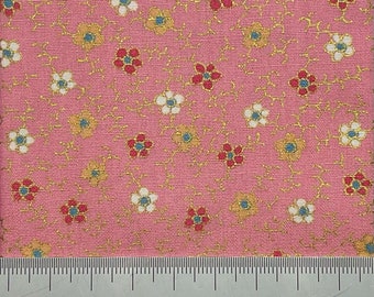 Op de afbeelding: Roze stof met een herhalend patroon van kleine witte, rode en blauwe bloemen met gouden accenten. Onderaan de afbeelding is een liniaal zichtbaar met metingen in inches.