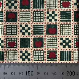 May include: A beige fabric with a repeating pattern of red apples, green squares, and black and white checkered squares. The fabric is measured with a ruler showing 150 cm.