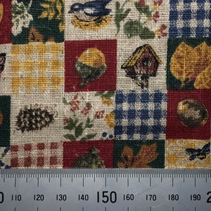 May include: A woven fabric featuring a patchwork grid of squares with various nature-themed prints including birdhouses, blue birds, pinecones, autumn leaves, and checkered patterns. A metal ruler is positioned at the bottom for scale.
