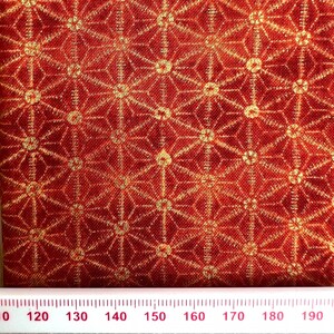 May include: Close-up of a red fabric with a repeating geometric pattern in gold. The pattern features star-like shapes and lines. A ruler with inch markings is at the bottom of the image.