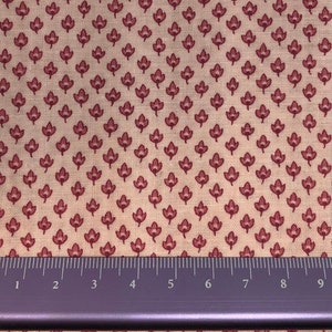 May include: A close-up of a fabric with a repeating pattern of small, red stylised leaves on a light beige background. A ruler is placed at the bottom of the image to show scale.