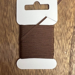 May include: A spool of brown cord on a white card with a hanging hole. The cord is wound tightly and has a textured surface.