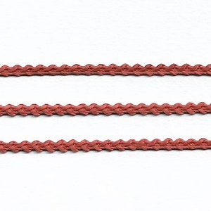 May include: Three strands of coral-colored decorative trim are displayed against a white background. The trim has a braided or looped design, creating a textured appearance. The color is a muted, earthy tone.