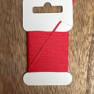 May include: A spool of red cord on a white card with a hanging hole. The cord is wound tightly and is ready to be used for crafts or other projects.