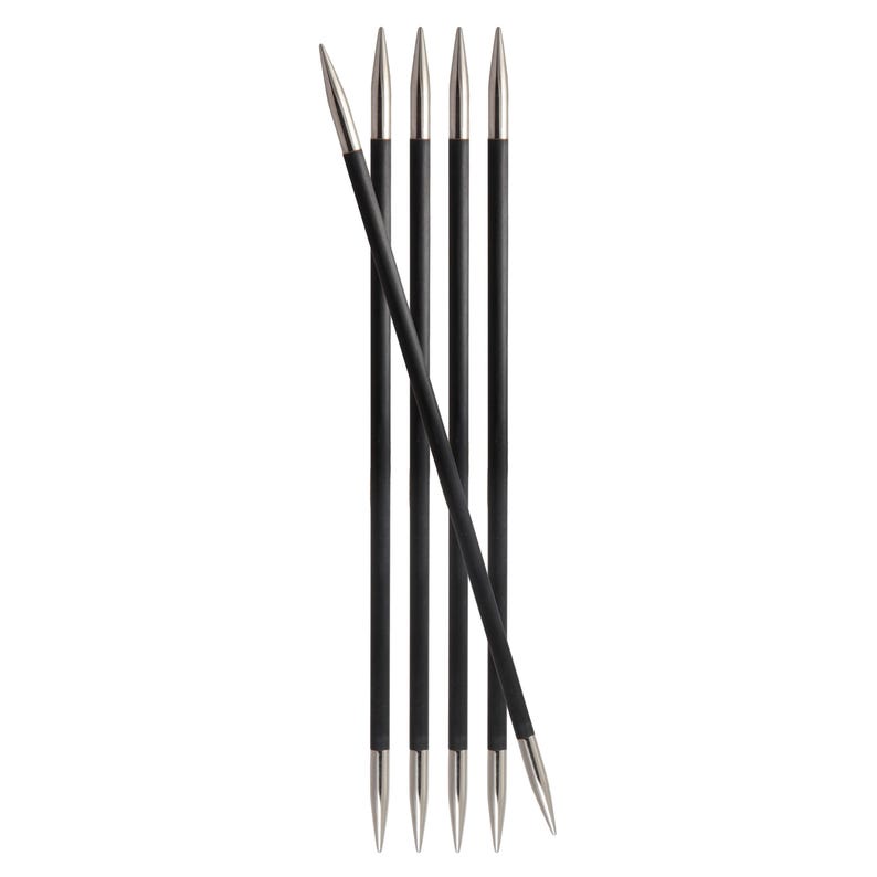 May include: A set of six knitting needles. The needles have black shafts and silver-colored tips. One needle is angled across the others. The needles are arranged vertically against a white background.