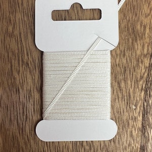 May include: A spool of off-white cord, neatly wound and attached to a white cardboard tag. The cord is thin and appears to be made of a woven material. The tag has a hole for hanging and a rounded bottom.