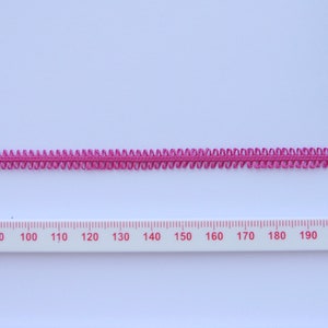 May include: A length of magenta-colored decorative trim with a scalloped edge. The trim is placed on a white surface next to a ruler marked in centimeters. The trim appears to be made of a woven material.