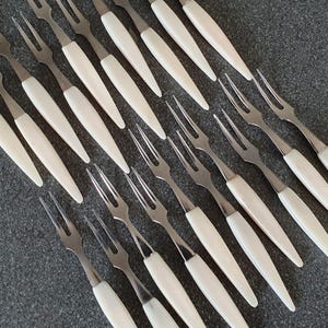Pode incluir: Um conjunto de dezasseis garfos de cocktail em aço inoxidável dispostos numa grelha. Cada garfo apresenta uma cabeça metálica de dois dentes e um cabo branco liso e cónico. Os garfos têm um design uniforme com acabamento prateado.