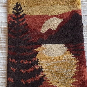 May include: A rectangular, textured wall hanging depicting a sunset over a mountain range. The artwork features a color palette of deep reds, browns, yellows, and white, with a detailed tree silhouette on the left side. The piece is likely handmade.
