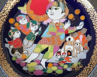 May include: A decorative plate with a dark blue background and a gold rim. The plate features a colourful illustration of a person juggling oranges, surrounded by children and buildings. The design has a whimsical, folk art style.