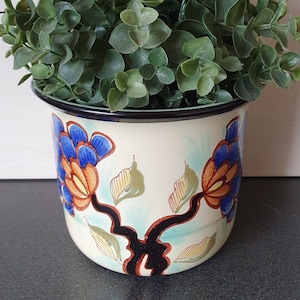 Może przedstawiać: Biała ceramiczna doniczka z niebieskim, pomarańczowym i zielonym wzorem kwiatowym. Doniczka jest wypełniona zieloną, liściastą rośliną.