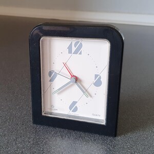 Puede incluir: Reloj despertador negro con una esfera blanca y números negros. El reloj tiene una aguja de segundos roja y una aguja de minutos y horas gris. La esfera del reloj también tiene líneas y números grises. El reloj dice "Quartz" y "G. Hellen Made in Germany" en la parte inferior.
