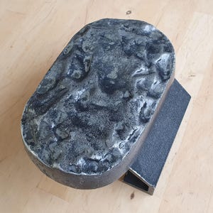 Puede incluir: Objeto metálico gris oscuro con una textura irregular y forma orgánica, montado sobre una base rectangular. La parte superior tiene una superficie rugosa y picada, mientras que la base es lisa y angular.