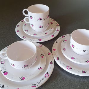 May include: A set of three white ceramic teacups and saucers with a geometric pattern in black and pink. The cups and saucers have a speckled finish.