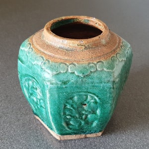 May include: A hexagonal ceramic jar with a green glaze and brown accents. The jar has a raised design of flowers and leaves.