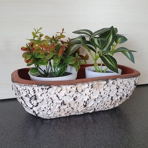 Op de afbeelding: Een ovale plantenbak met een getextureerde wit en zwarte buitenkant en een bruine binnenkant. Het bevat twee kleine potplanten met groene en rode bladeren. De plantenbak staat op een donkere ondergrond.