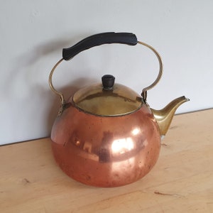 Puede incluir: Una tetera de cobre con tapa de latón y asa de plástico negro. La tetera tiene un cuerpo redondeado y un pico en el lateral.