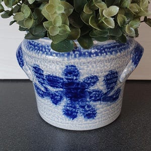 May include: A blue and white ceramic pot with a floral design. The pot has a textured surface and two small handles. The pot is filled with green leafy plants. The pot is likely used for decorative purposes.