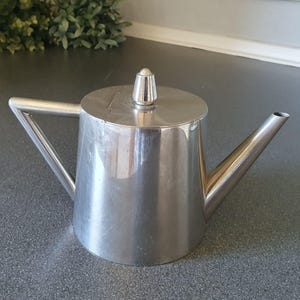 Op de afbeelding: Een theepot van gepolijst zilverkleurig metaal met een conisch lichaam, een plat deksel met een kleine puntige knop, een driehoekig handvat en een rechte, schuine tuit. De theepot heeft een glad, reflecterend oppervlak en een strak ontwerp.