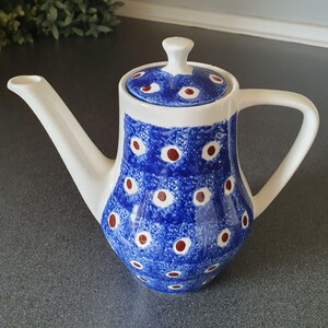 Peut inclure: Théière en céramique blanche avec un motif bleu et blanc de formes en forme d'yeux et de points rouges. La théière a un bec courbé, une anse et un couvercle avec un petit bouton. Le motif est répété sur le couvercle.