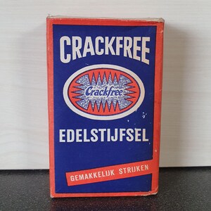 Pode incluir: Uma caixa vermelha e azul com o texto "Crackfree Edelstijfsel" e "Gemakkelijk strijken". A caixa tem um oval vermelho com uma estrela branca e o texto "Crackfree" em branco.