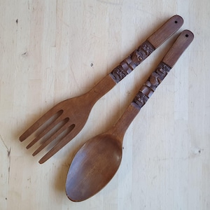 May include: A wooden fork and spoon set. The utensils are made of dark brown wood and feature carved details on the handles. The fork has four tines, and the spoon has a rounded bowl. The set is displayed on a light wood surface.