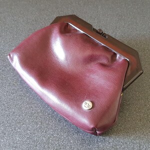 Può includere: Una pochette bordeaux con chiusura marrone scuro. La piccola borsa ha un bottone argentato con un emblema circolare. Il materiale simile alla pelle è leggermente strutturato.