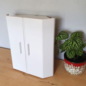 May include: White corner cabinet with two doors and two handles. The cabinet is made of plastic and has a simple design.