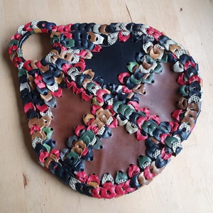 Puede incluir: Un bolso redondo de cuero marrón con un diseño único. El bolso está hecho de pequeñas piezas de cuero de colores que están cosidas juntas en un patrón. El bolso tiene un asa y un cierre de solapa.