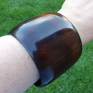 Op de afbeelding: Een brede, donkerbruine armband met een glanzende afwerking. De armband is gebogen en wikkelt zich om de pols. De kleur is een rijk, warm bruin en het oppervlak reflecteert licht.